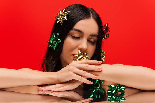 Young woman with makeup and gift bows on hair touching hand near reflective surface isolated on red  - Foto, imagen