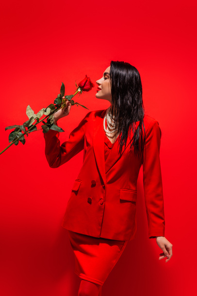 Side view of brunette woman in jacket and necklace smelling rose on red background  - Foto, Bild