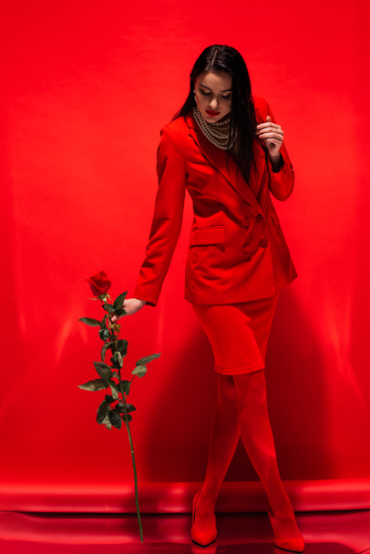Full length of stylish woman in jacket and pearl necklace holding rose on red background  - Фото, зображення
