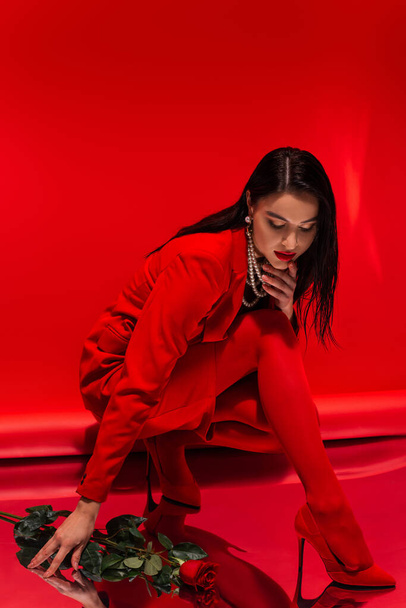 Stylish model in heels and pearl necklace looking at reflective surface near rose on red background  - 写真・画像