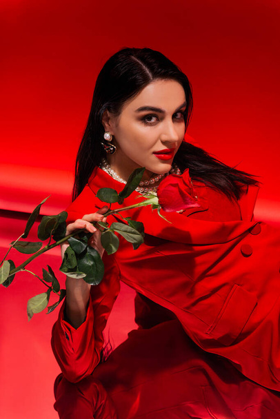 Pretty brunette woman in jacket holding rose and looking at camera on red background  - Photo, Image