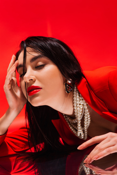 Портрет элегантной брюнетки в жемчужном ожерелье, лежащей на отражающей поверхности, изолированной на красном  - Фото, изображение