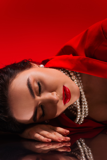 Young woman in red jacket and pearl necklace lying on reflective surface on red background  - Foto, Imagen