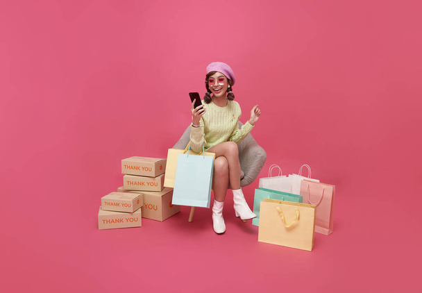 Wesoły szczęśliwy zakupoholiczka azjatycka kobieta z dużą ilością toreb na zakupy, siedzi na fotelu i za pomocą telefonu komórkowego, puste miejsce do kopiowania. - Zdjęcie, obraz