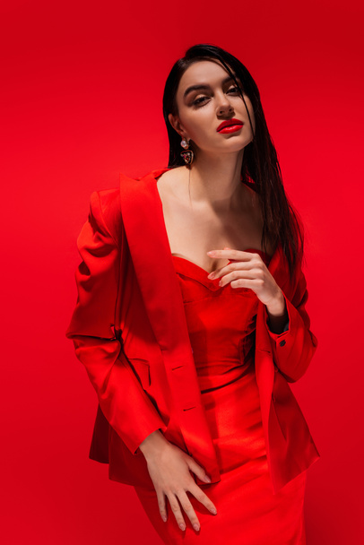 Pretty and elegant brunette model in jacket posing and looking at camera isolated on red  - Photo, Image