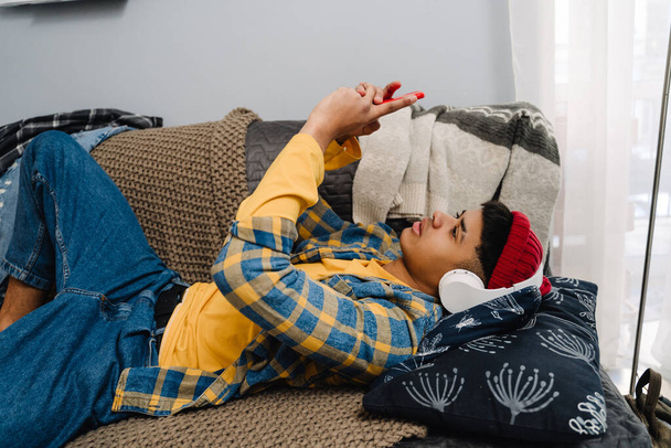 Middle-eastern teenage boy using headphones and cellphone while lying on couch at home - Foto, immagini