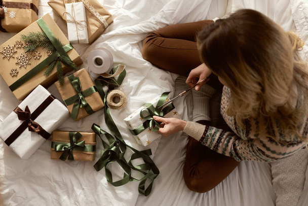Draufsicht einer kaukasischen Frau, die zu Hause auf dem Bett sitzt und Weihnachtsgeschenk einpackt  - Foto, Bild