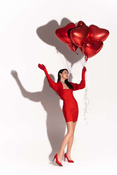 Cheerful woman in heels and dress holding red heart shaped balloons and waving hand on white background with shadow  - Photo, Image