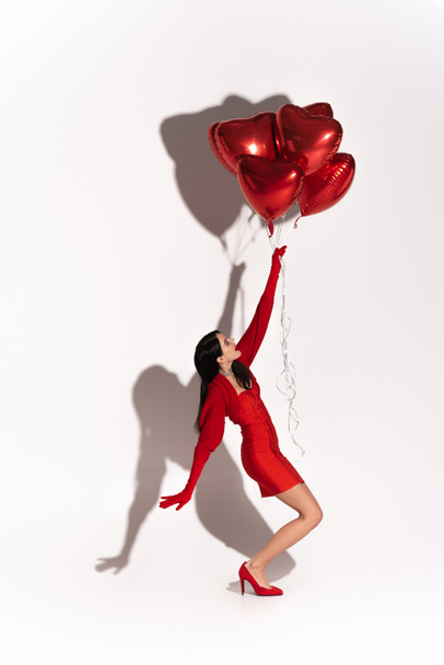 Side view of stylish woman in dress and heels pulling heart shaped balloons on white background with shadow  - Foto, immagini