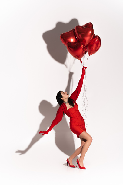 Side view of stylish model in dress looking at heart shaped balloons on white background with shadow  - Φωτογραφία, εικόνα