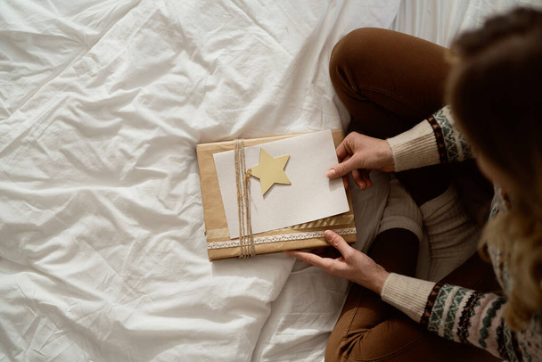 Draufsicht einer kaukasischen Frau, die auf dem Bett sitzt und Weihnachtsgeschenk mit Grußkarte verpackt  - Foto, Bild