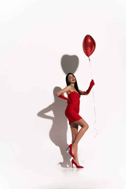Full length of sexy woman in red dress and gloves holding heart shaped balloon on white background with shadow  - Фото, зображення