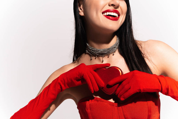 Καλλιεργημένη άποψη της χαρούμενης γυναίκας σε κόκκινα γάντια κρατώντας καρδιά σχήμα δώρο που απομονώνονται σε λευκό  - Φωτογραφία, εικόνα
