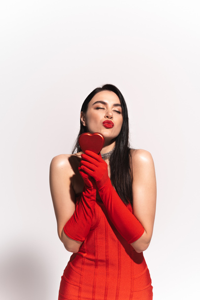 Stylish model in red dress and gloves holding heart shaped gift box and blowing air kiss on grey background  - Fotografie, Obrázek