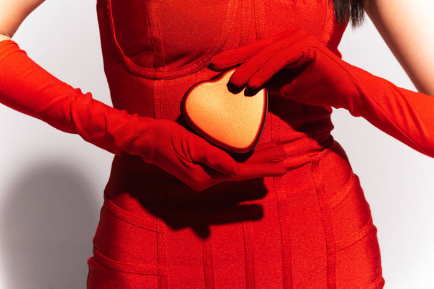 Cropped view of woman in red gloves and dress holding present in heart shape on grey background  - Photo, Image