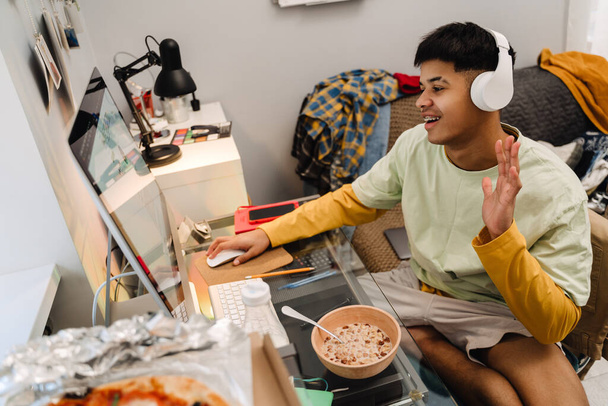 Brunette teenage boy talking on video call using desktop computer while having breakfast in bedroom - Foto, immagini