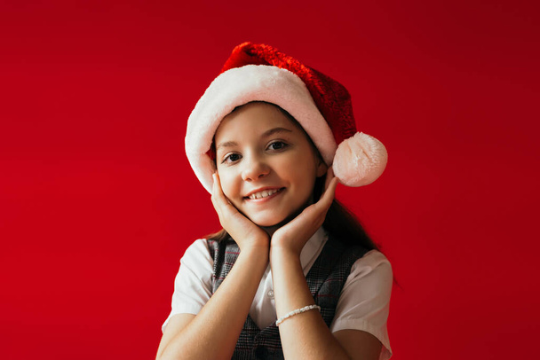smiling girl in santa hat and beaded bracelet holding hands near face isolated on red - Foto, Imagen