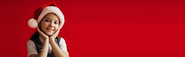 gioiosa ragazza in cappello di Babbo Natale sorridente alla fotocamera mentre si tiene le mani vicino al viso isolato sul rosso, banner - Foto, immagini