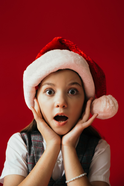 Porträt eines erstaunten Mädchens mit Weihnachtsmütze, das in die Kamera blickt und die Hände in der Nähe des Gesichts hält, isoliert auf rotem Grund - Foto, Bild