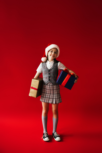 teljes hossza trendi és boldog lány télapó kalap és kockás ruhák gazdaság ajándék dobozok piros háttér - Fotó, kép
