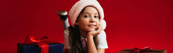 chica feliz en sombrero de santa que yace cerca de regalos de Navidad mientras mira la cámara aislada en rojo, pancarta - Foto, Imagen