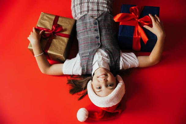 top view of joyful girl in plaid vest and santa hat lying near gift boxes on red background - Fotografie, Obrázek