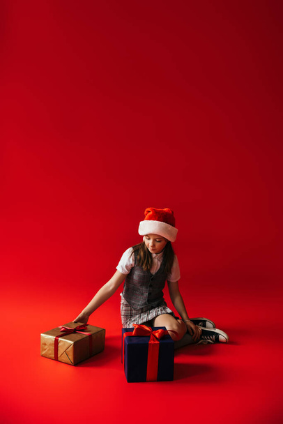 full length of preteen girl in santa hat and gumshoes sitting near gift boxes on red background with copy space - Photo, image