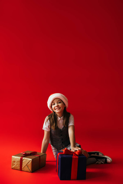 iloinen tyttö santa hattu istuu lähellä uutta vuotta esittelee punaisella taustalla kopioi tilaa - Valokuva, kuva