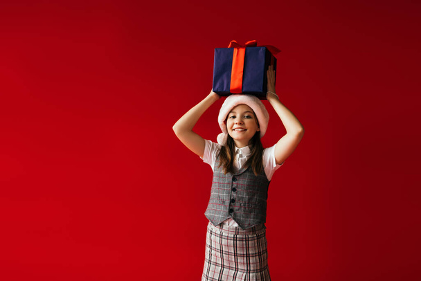 cheerful girl in plaid clothes and santa hat holding Christmas presents above head isolated on red - 写真・画像