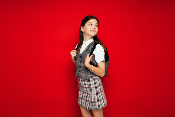 χαμογελαστό κορίτσι με καρό ρούχα στέκεται με σακίδιο και κοιτάζοντας πίσω απομονώνονται σε κόκκινο, banner  - Φωτογραφία, εικόνα
