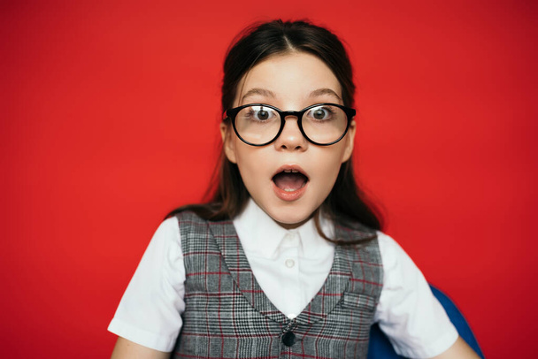 astonished girl in eyeglasses and plaid vest looking at camera isolated on red - Photo, Image