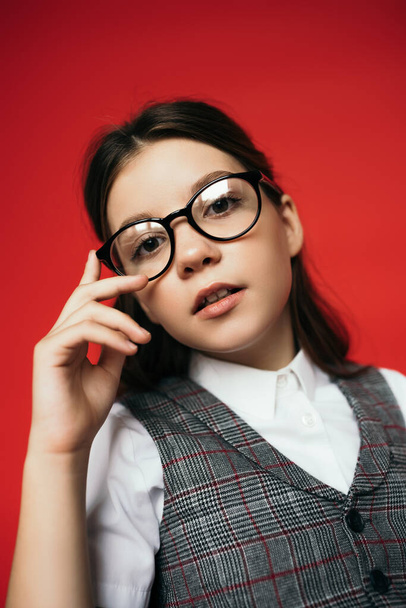 portrait of preteen girl in trendy eyeglasses posing and looking at camera isolated on red - Foto, Imagen