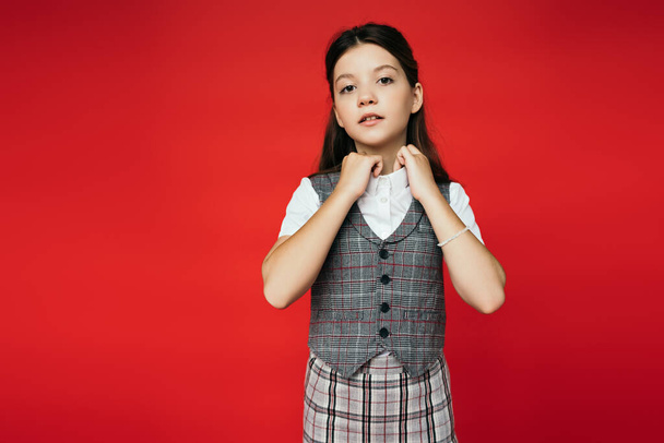 preteen girl in checkered vest and skirt adjusting collar of white blouse isolated on red, banner  - Fotoğraf, Görsel