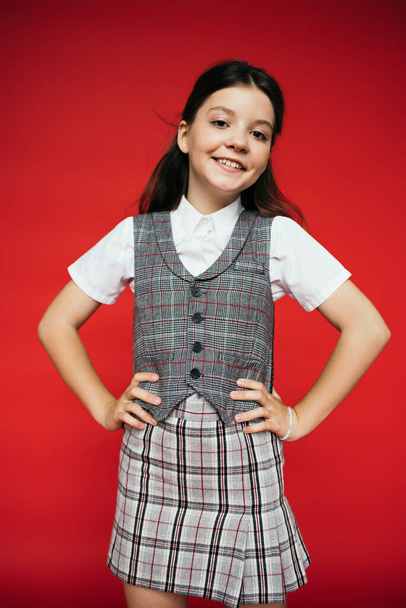 joyful girl in checkered skirt and vest standing akimbo and looking at camera isolated on red, banner  - Foto, Imagen