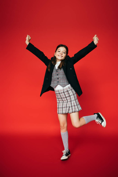 full length of excited girl in plaid skirt and black blazer posing on one leg with outstretched hands on red background - Foto, afbeelding
