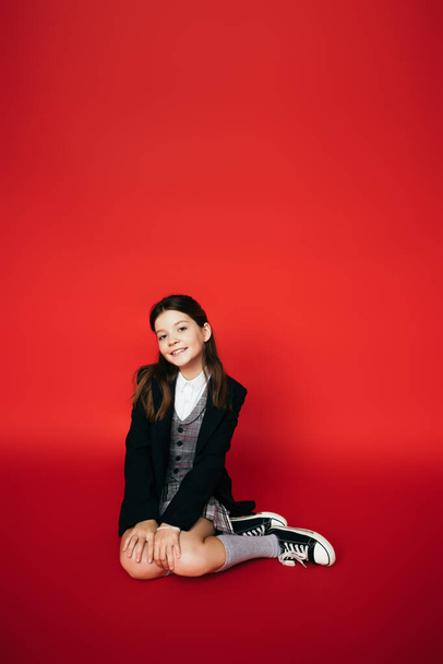full length of stylish girl in gumshoes and black blazer smiling at camera while sitting on red background - Fotografie, Obrázek