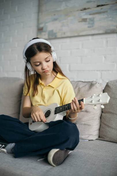 displeased girl in headphones playing little hawaiian guitar while sitting on couch with crossed legs - Fotografie, Obrázek