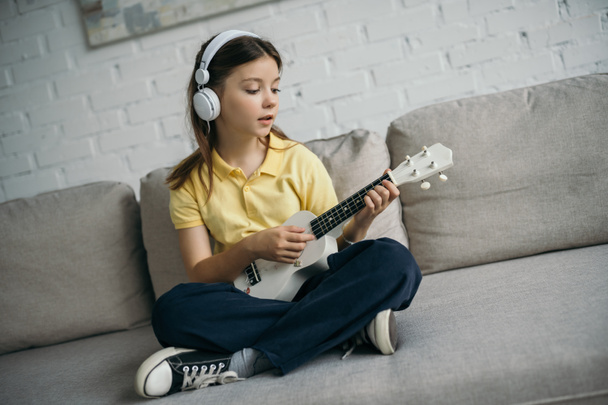girl in wired headphones sitting on sofa with crossed legs and playing ukulele - Zdjęcie, obraz