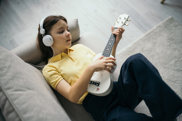 top view of girl in wired headphones lying on sofa and playing small hawaiian guitar - Fotografie, Obrázek