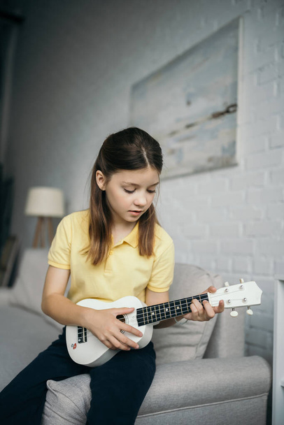 preteen girl sitting on couch in blurred living room and playing ukulele - Foto, imagen