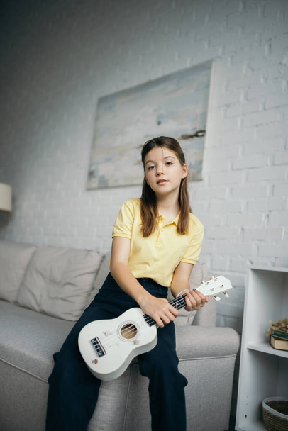 preteen girl with small hawaiian guitar looking at camera near couch in living room - Фото, изображение