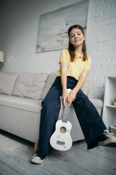 pleased preteen girl with little hawaiian guitar looking at camera near couch in living room - Fotografie, Obrázek