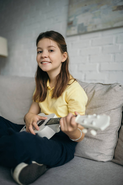 cheerful preteen girl smiling at camera while playing ukulele on sofa in living room - Foto, Imagem