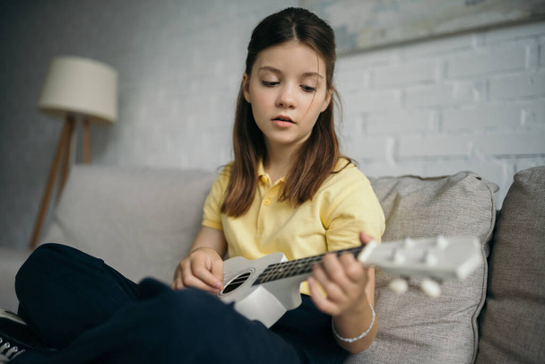 brunette girl playing ukulele while sitting on sofa at home - 写真・画像