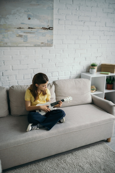 wysoki kąt widzenia dziewczyny siedzi na nowoczesnej kanapie ze skrzyżowanymi nogami i gra ukulele - Zdjęcie, obraz