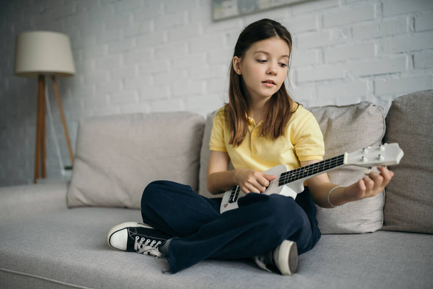 брюнетка дівчина сидить на дивані з схрещеними ногами і налаштовує маленьку гавайську гітару
 - Фото, зображення