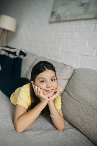 alegre chica escuchando música en auriculares inalámbricos mientras está acostado en el sofá con las manos cerca de la cara - Foto, imagen