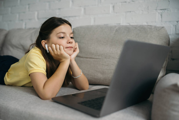 fille brune dans un écouteur sans fil regardant l'ordinateur portable flou tout en étant allongé sur un canapé confortable à la maison - Photo, image