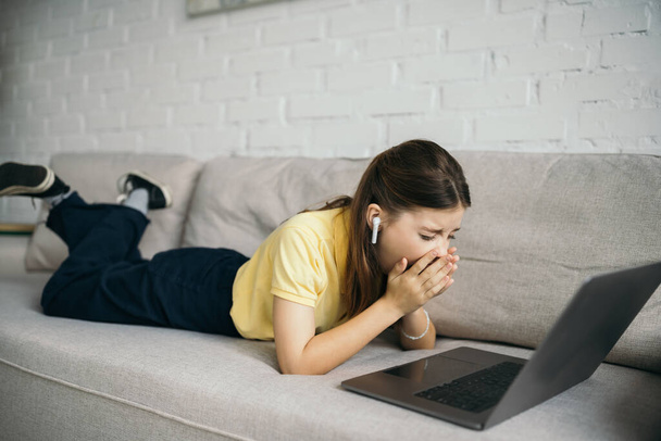bored girl in wireless earphone yawning near laptop on comfortable couch in living room - Photo, image