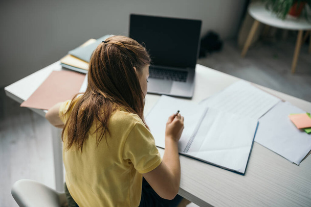 hoge hoek uitzicht van brunette meisje schrijven in notebook in de buurt laptop met blanco scherm thuis - Foto, afbeelding
